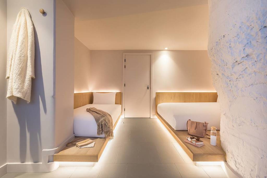 An ultra modern, yet small, hotel room. Twi twin beds with recessed lighting both in the ceiling, on the walls, and under the frames of the beds illuminates the room with elegance. Clean white plain walls, modern wood, and slate flooring make an ultra-clean, super modern, small room.