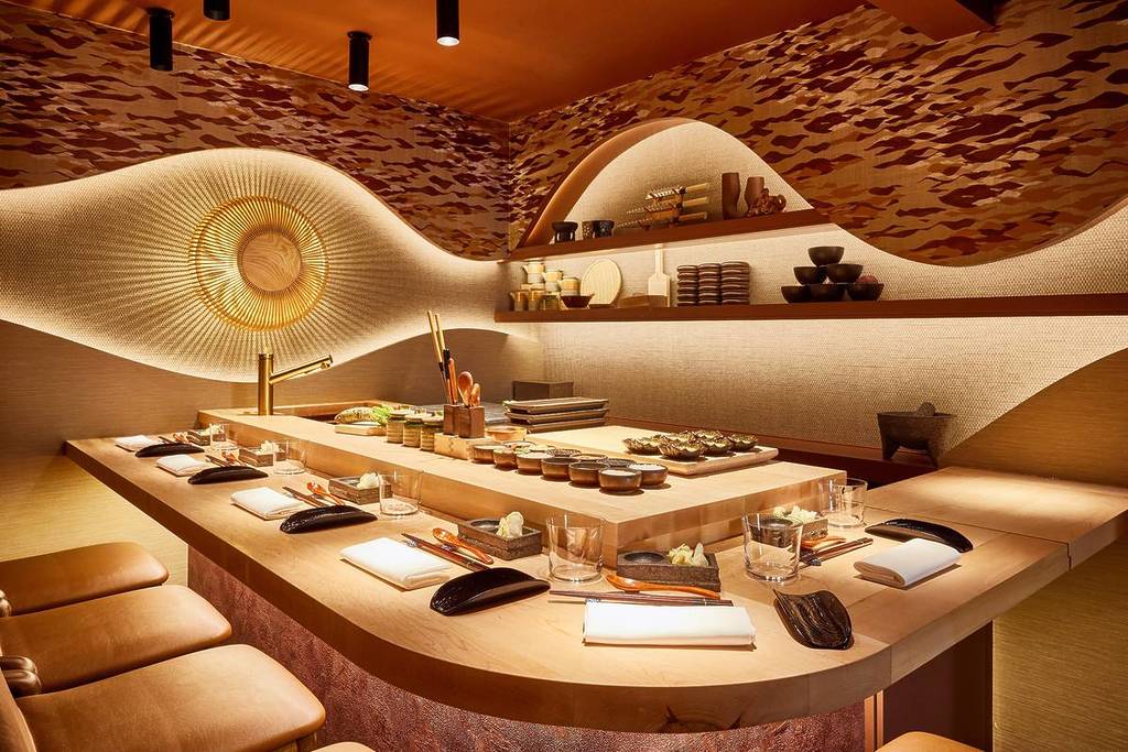 L'image montre un bar à sushis avec un long comptoir en bois clair. Le comptoir est aménagé pour les convives avec des couverts comprenant des baguettes, des petits plats et des verres. Derrière le comptoir se trouve une zone de préparation avec divers ingrédients et outils, ainsi que des étagères contenant de la vaisselle et des ustensiles supplémentaires. Les murs sont décorés d'un matériau texturé de couleur terre et d'une grande décoration murale en forme de soleil doré.