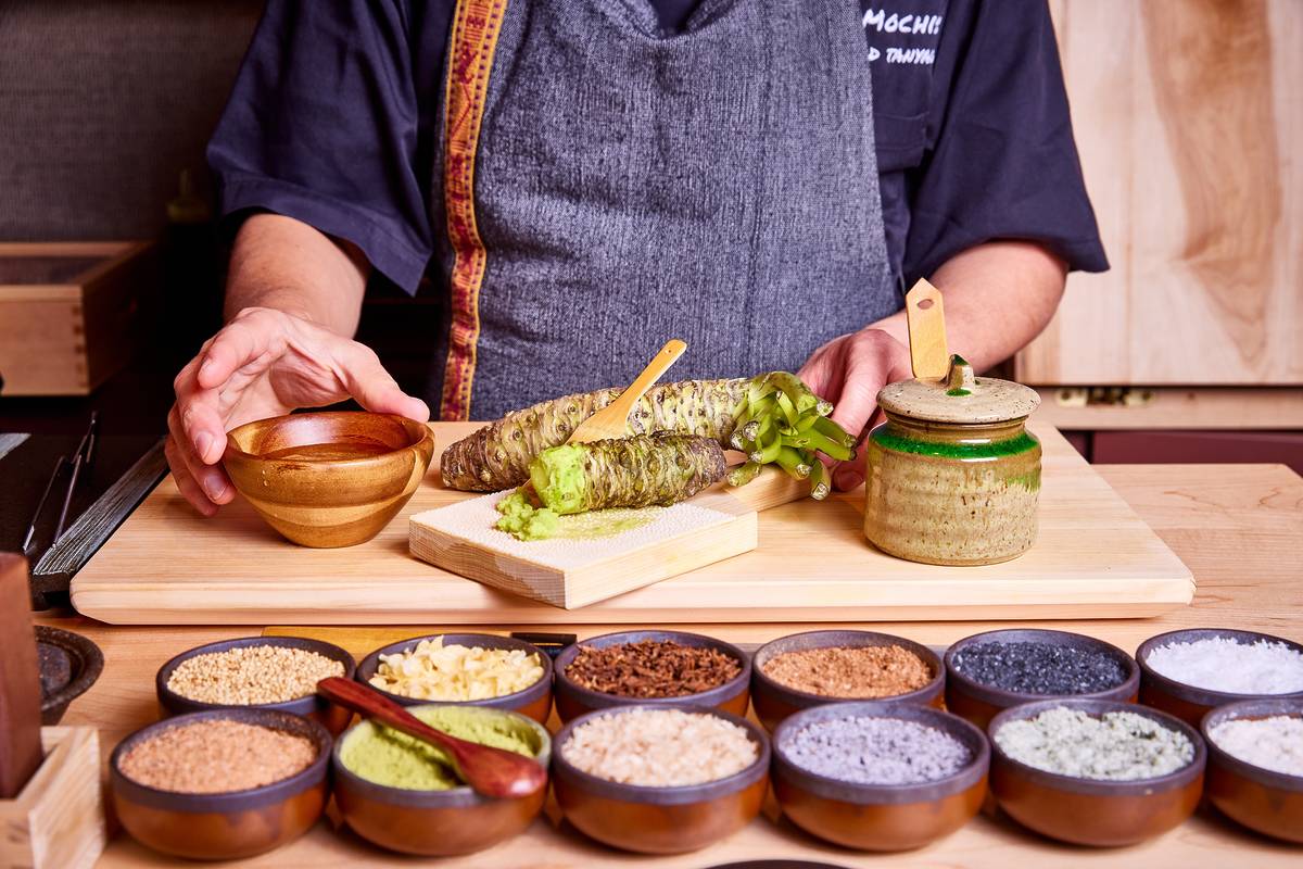 The image shows a chef preparing wasabi. He is grating fresh wasabi root onto a wooden board. A small wooden bowl, a small ceramic jar with a lid, and several small bowls containing various spices and seasonings are also visible on the counter.