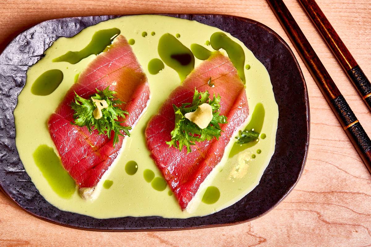 Two pieces of what appears to be seared tuna are artistically arranged on a dark-colored, textured plate. The tuna rests atop a pale green sauce or purée, with small droplets of a darker green oil or dressing artfully scattered across the surfac