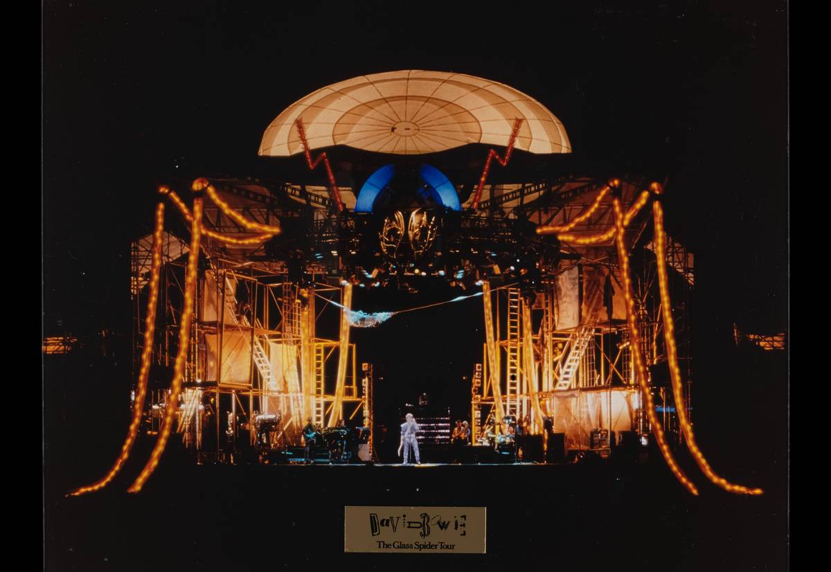 A dramatic nighttime concert stage setup from David Bowie's 1987 Glass Spider Tour. The stage features a massive spider-like structure with glowing orange legs made of lights extending from a central platform. At the center, under a large domed canopy, David Bowie stands illuminated in a sparkling outfit. The background is filled with intricate scaffolding, ladders, and theatrical lighting, creating a surreal and futuristic atmosphere.