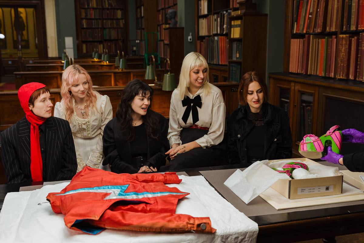 Five women, members of the UK band The Last Dinner Party, sit together in a historic library room, looking engaged as they observe iconic fashion items from David Bowie’s archive. In front of them on a table is a red stage outfit with a blue lightning bolt, and to the right, a conservator wearing purple gloves holds up a pair of platform shoes in bright pink and green. The room is lined with wooden bookshelves filled with vintage books, creating a rich, academic atmosphere.