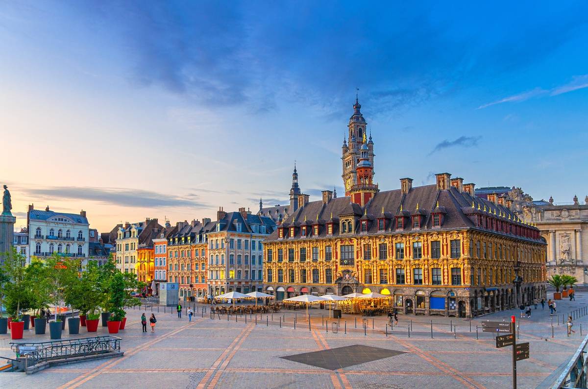 A large, open square in Lille, France, surrounded by old 17th-century buildings with colorful exteriors and steep roofs. In the middle's the Old Stock Exchange (La Vieille Bourse), a poshly decorated Renaissance building with red and gold details. Behind it's a tall clock tower with fancy stonework and a spire. People walk through the square, some perching at a café with white umbrellas. The sky's blue and orange, maybe early evening.