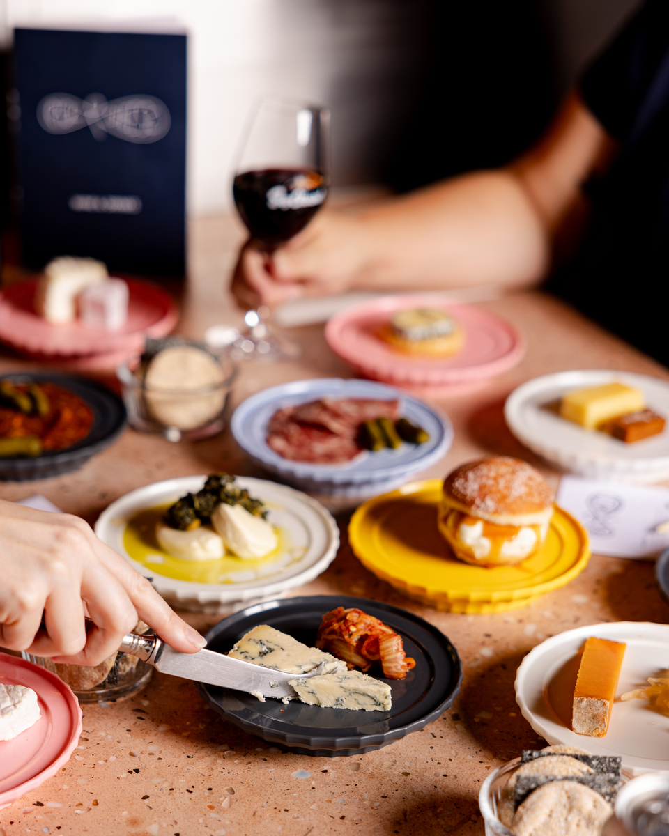 people digging in and cutting pieces of cheese from an array of colourful plates topped with cheese and cheese forward dishes