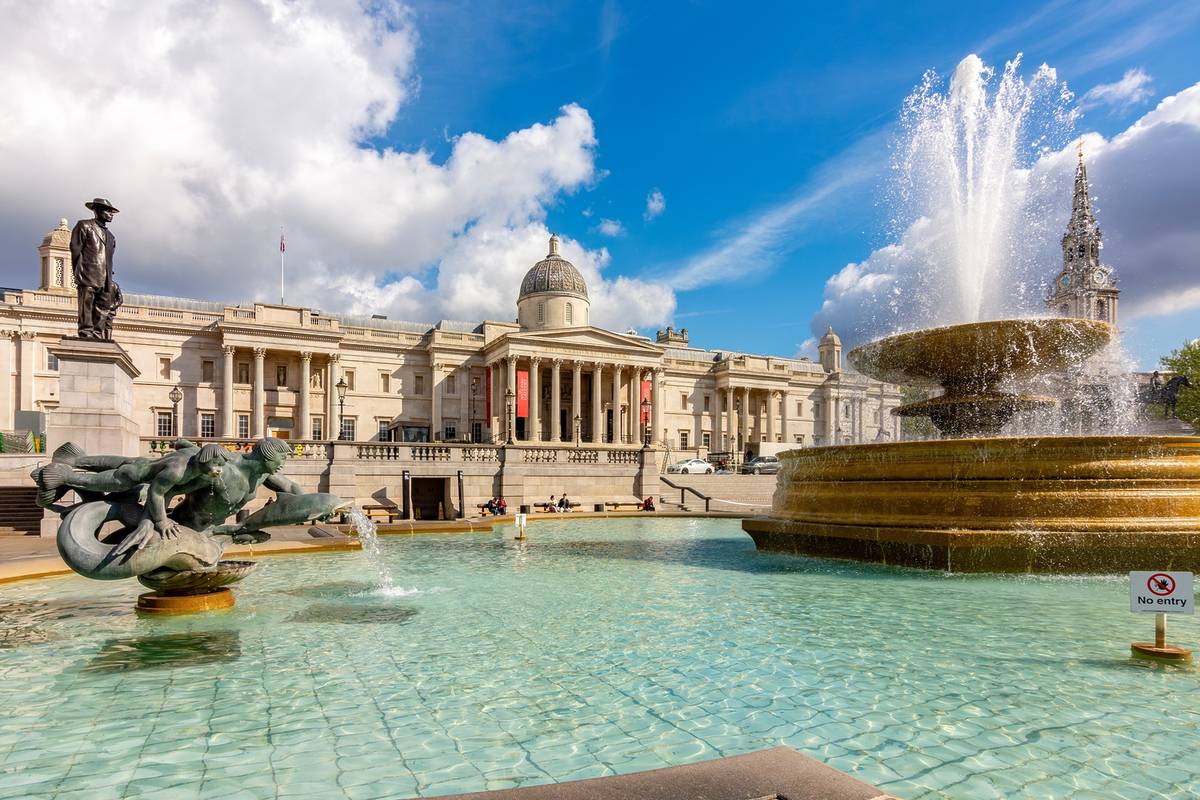 Exterior of The National Portrait Gallery in Trafalgar Square. Foreground's a blue watered fountain, and the museum stands elegantly in the background.
