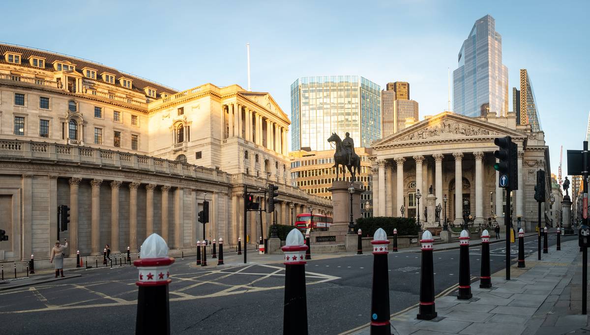 **Alt text:** A wide-angle view of the Bank of England Museum area in London, featuring the neoclassical Bank of England building on the left and the Royal Exchange on the right. A statue of the Duke of Wellington on horseback stands prominently in the center. Modern skyscrapers rise in the background, contrasting with the historic architecture. The street in the foreground is lined with distinctive black bollards topped with white domes and red stars.