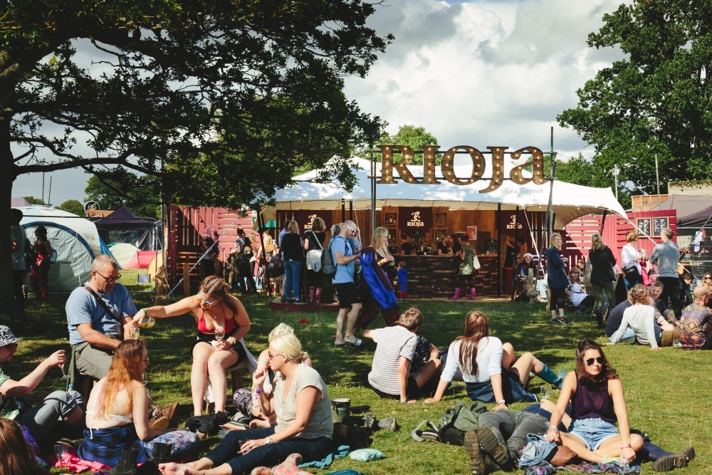 Groups of friends chilling on the grass outside a festival tent. A sign reads 'Rioja' above the tent's entrance.