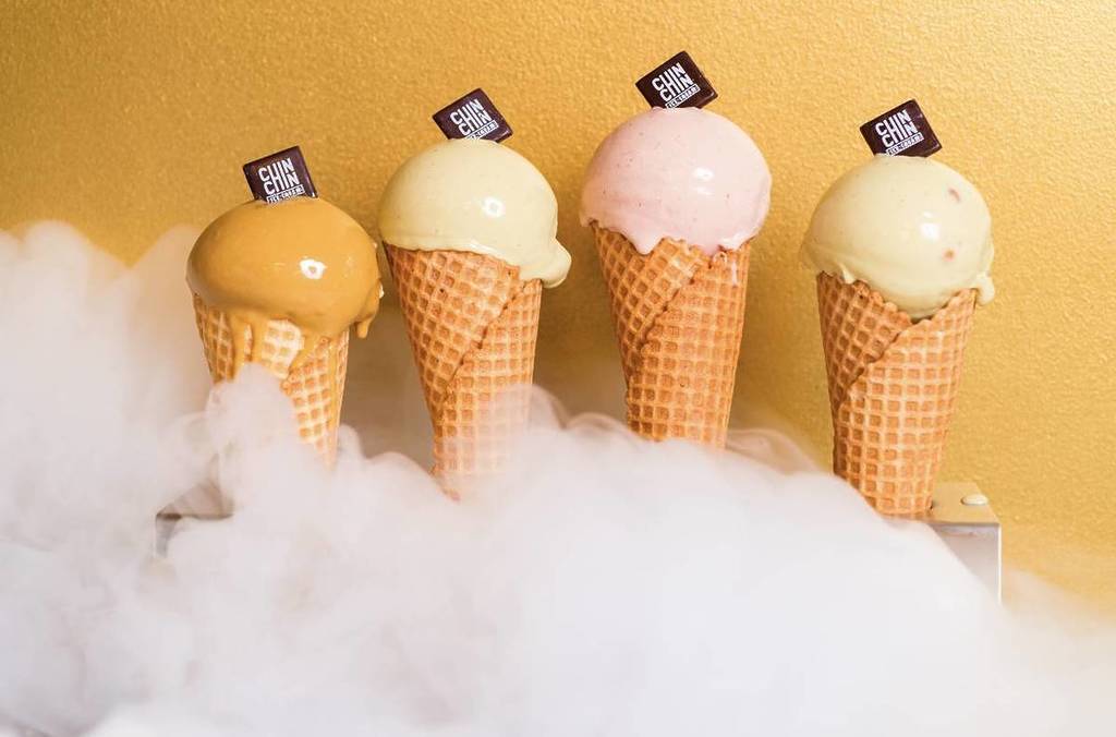 vier wafelhoorntjes die gedeeltelijk bedekt zijn door vloeibare stikstof, met glanzende bolletjes ijs, elk met een chocolaatje in het bolletje ijs met de tekst 'Chin Chin'.