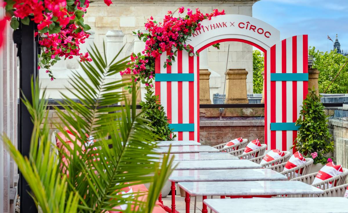A vibrant rooftop terrace at Kitty Hawk Restaurant in London, styled vibrantly. The space features red-and-white striped archways adorned with lush pink bougainvillaea, marble-top tables, and red-accented seating. Green potted plants line the area, and a grand historic stone building provides a striking backdrop under a bright blue sky.