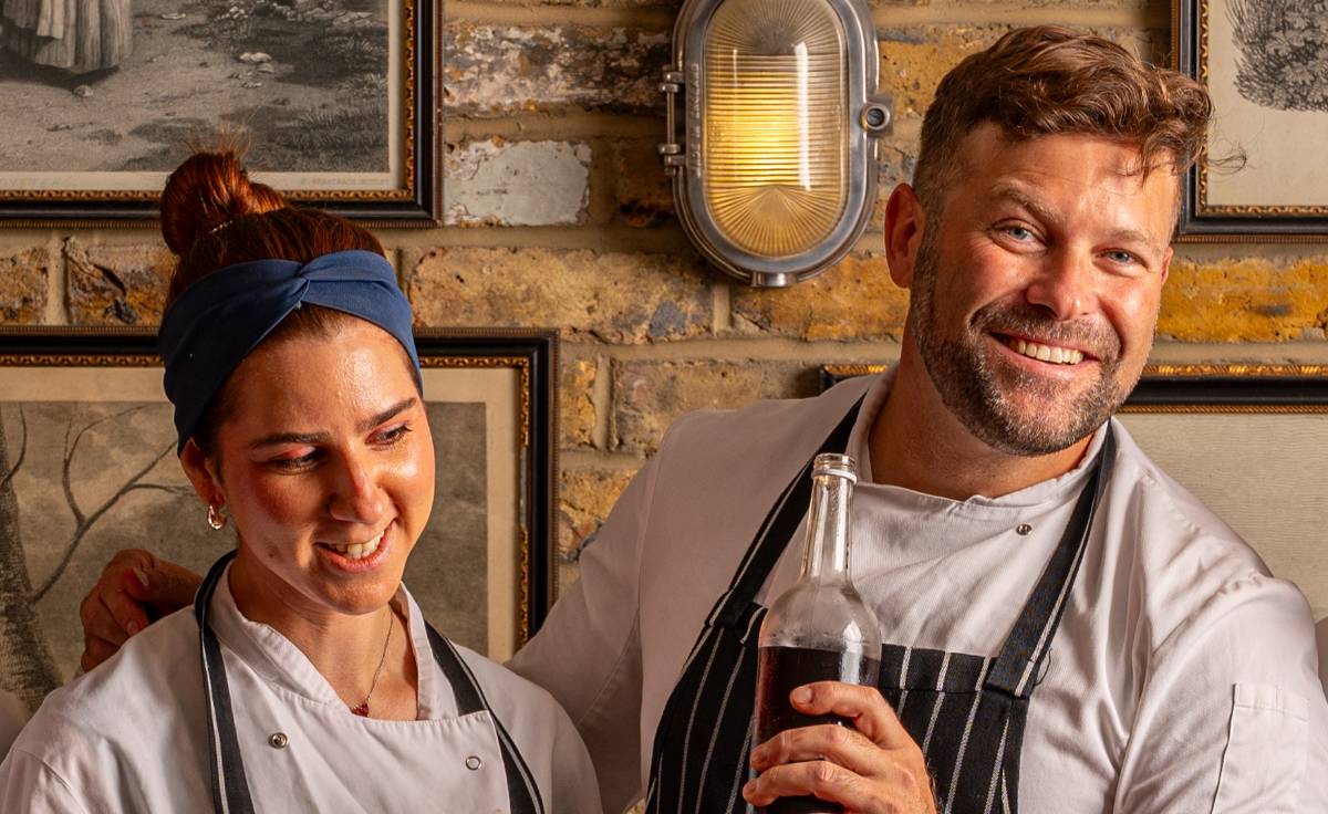 two chefs smile warmly, in front of a brick-walled background. the foreground shows a brick wall with photo frames. one chef on the right is smiling, while the one of the left is looking off-camera with a grin.
