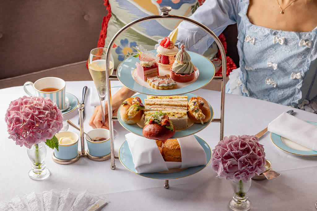 une personne assise, vêtue d'une robe à l'allure de Marie-Antoinette, derrière un plateau à trois étages garni de délicieux et adorables mets sucrés et salés, entourés de scones, de coupes de champagne et de tasses de thé.