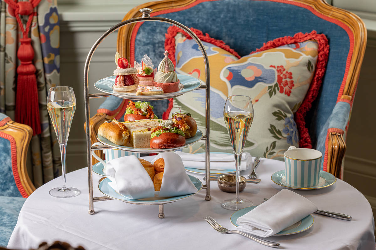 an afternoon tea spread of delectable and adorable looking sweet and savoury delights on a three tier tray, surrounded by scones, glasses of champagne, and mugs of tea