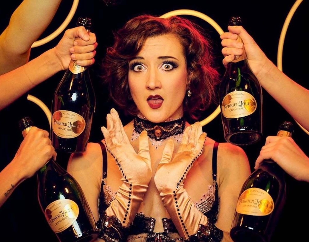 Sabrage acctress dressed in fancy dress with gloves surrounded by bottles of bubbly