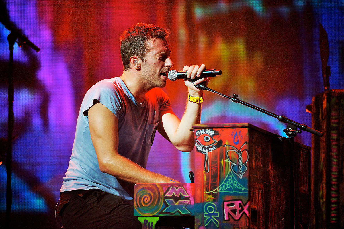 Chris Martin from the band Coldplay sat at a colourfully painted and decorated piano playing with one hand while singing into a microphone held by the other hand