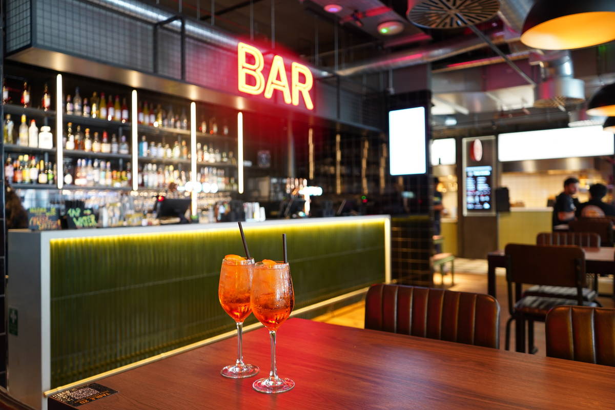 two aperol spritzes on a table in a bar, with a neon sign overheard reading 'bar'