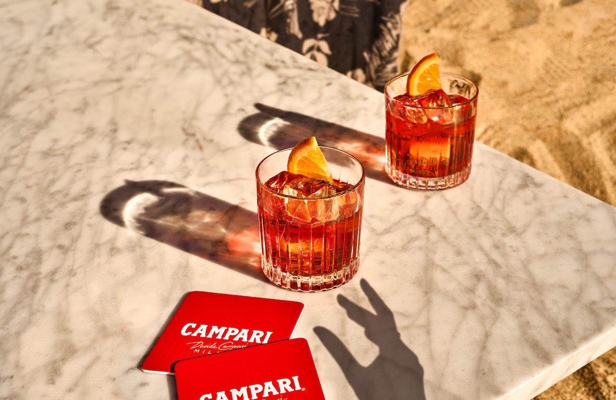 two negronis glowing in the sunshine on a table, next to two campari coasters