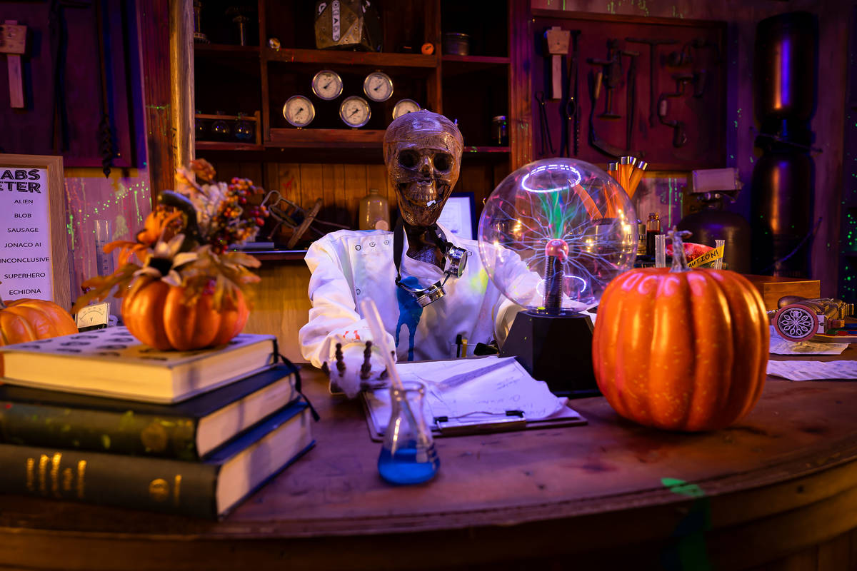 a spooky skeleton figurine sat at a desk in a lab coat surrounded by pumpkins and mysterious science-y gadgets