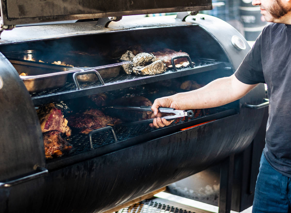 a tong-weielding hand reaching into a large smoker to move meats around on the shelves