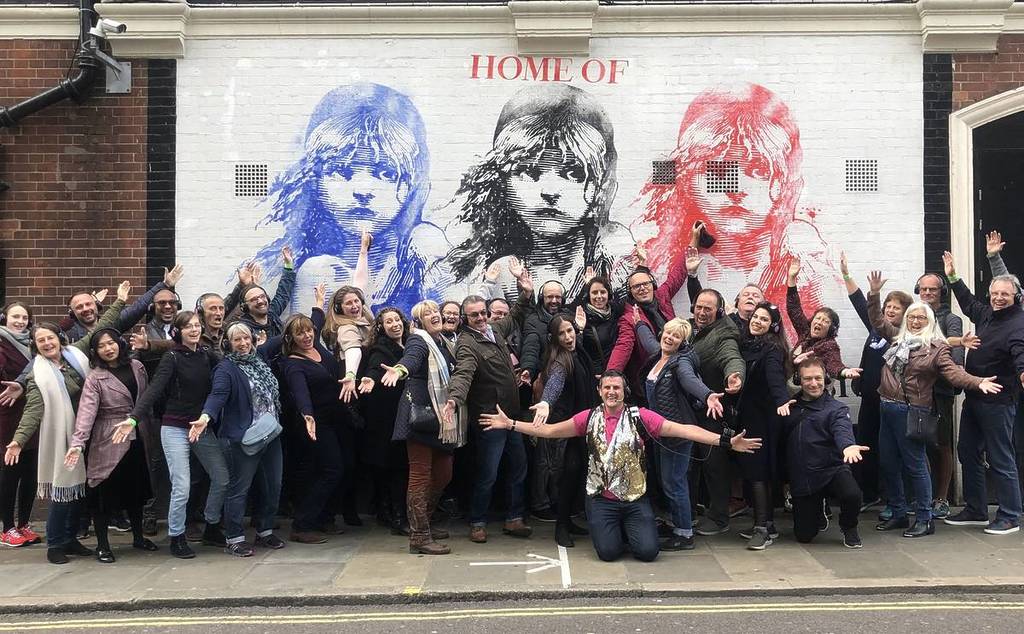 A walking tour group with silent disco headsets on posing in front of the Les Mis theatre sign