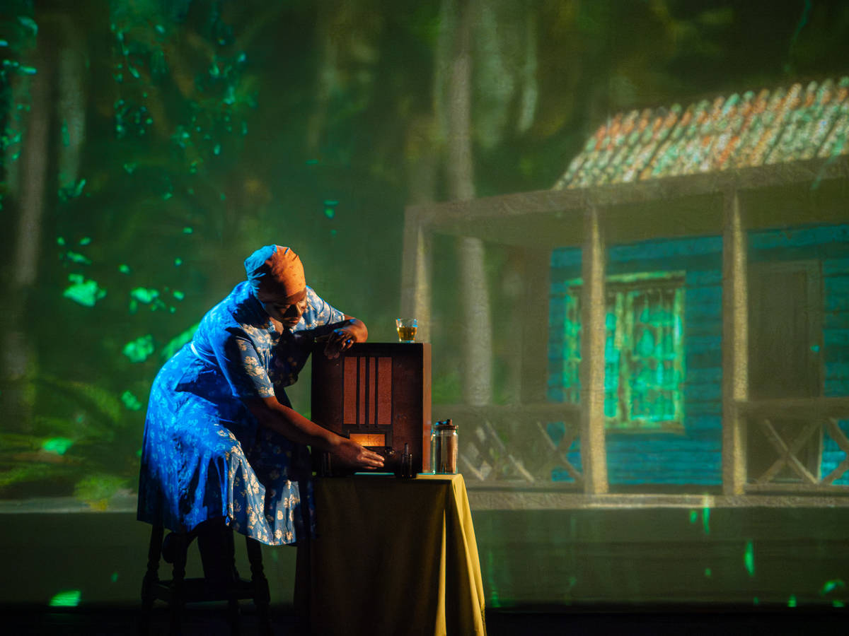 an actor onstage, portraying a Black woman in Barbados, interacts with a radio