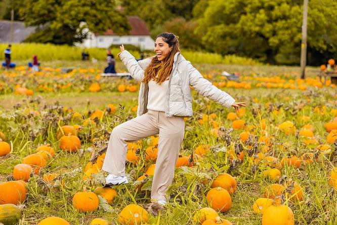 A lady stand in a field of pumpkins with her arms wide and a big smile on her face.