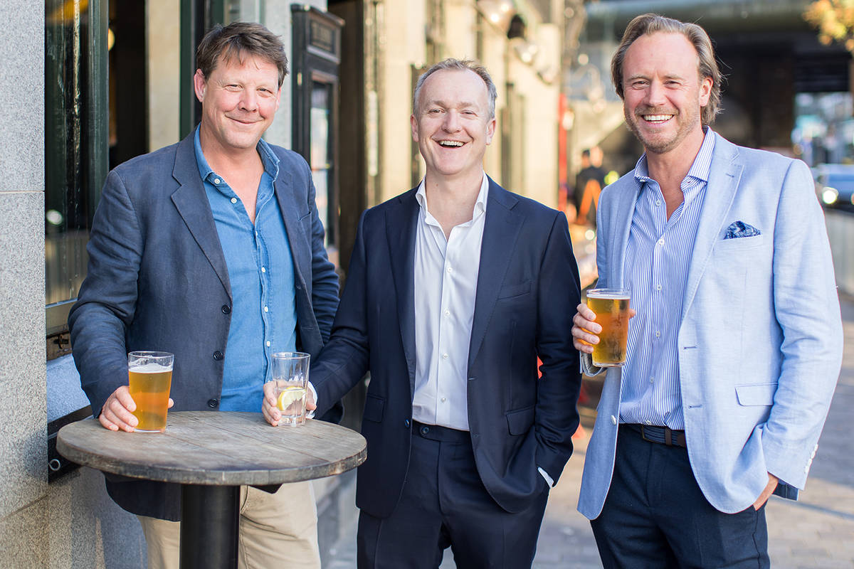 Three people stood side by side holding drinks outside a London pub