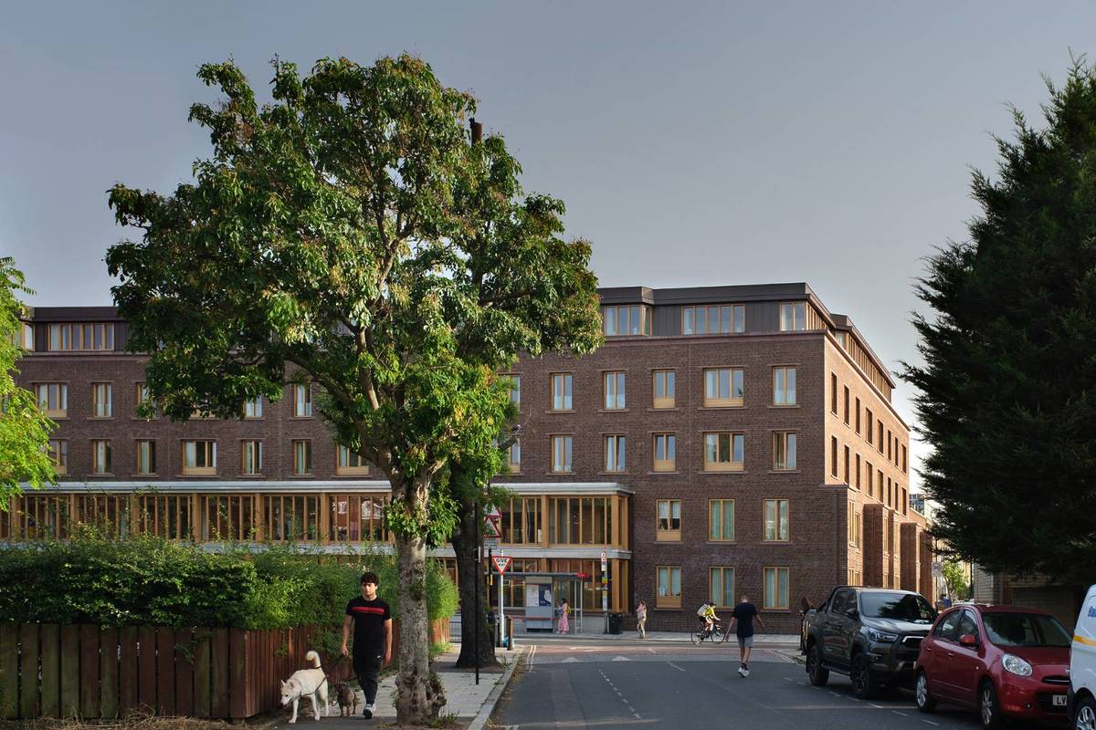 The exterior of a brick building with many windows on a street with trees, a bus stop and a man walking his dog.