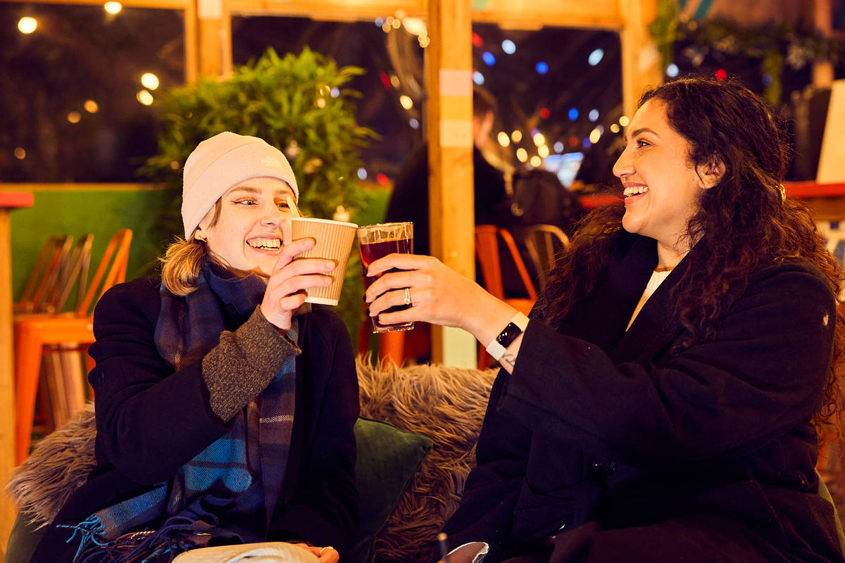 two people enjoying warm drinks and cocktails in a rooftop bar in winter, wrapped up in jackets and beanies