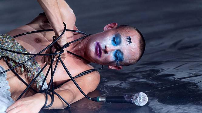 A performer in drag lying on the stage, tied up in a microphone cord with eyes closed