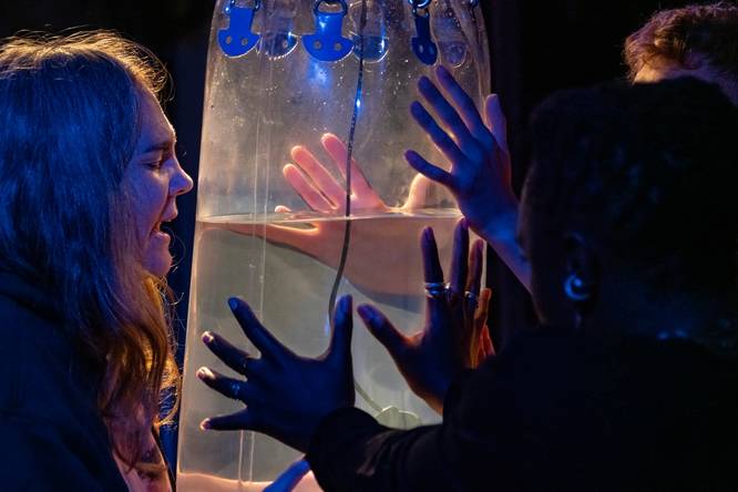 A theatre performer facing and singing at a large, water-filled boxing bag held up by two sets of hands