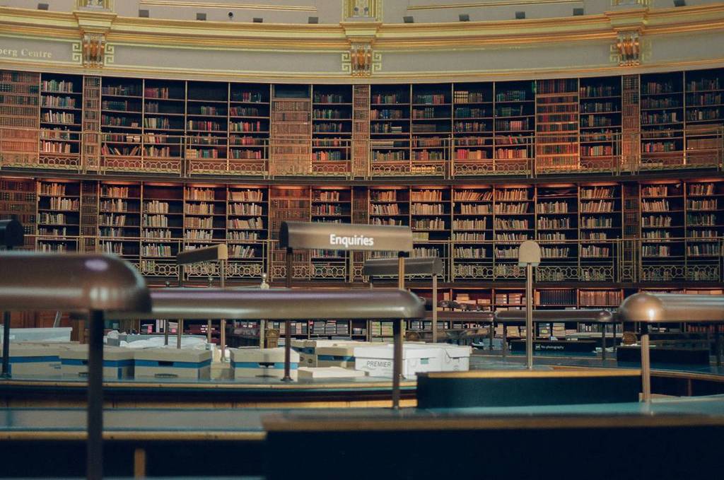 Reihenweise Bücher in einer riesigen Bibliothek mit goldenen Lichtern an der Decke und einem Bibliothekstisch im Vordergrund.