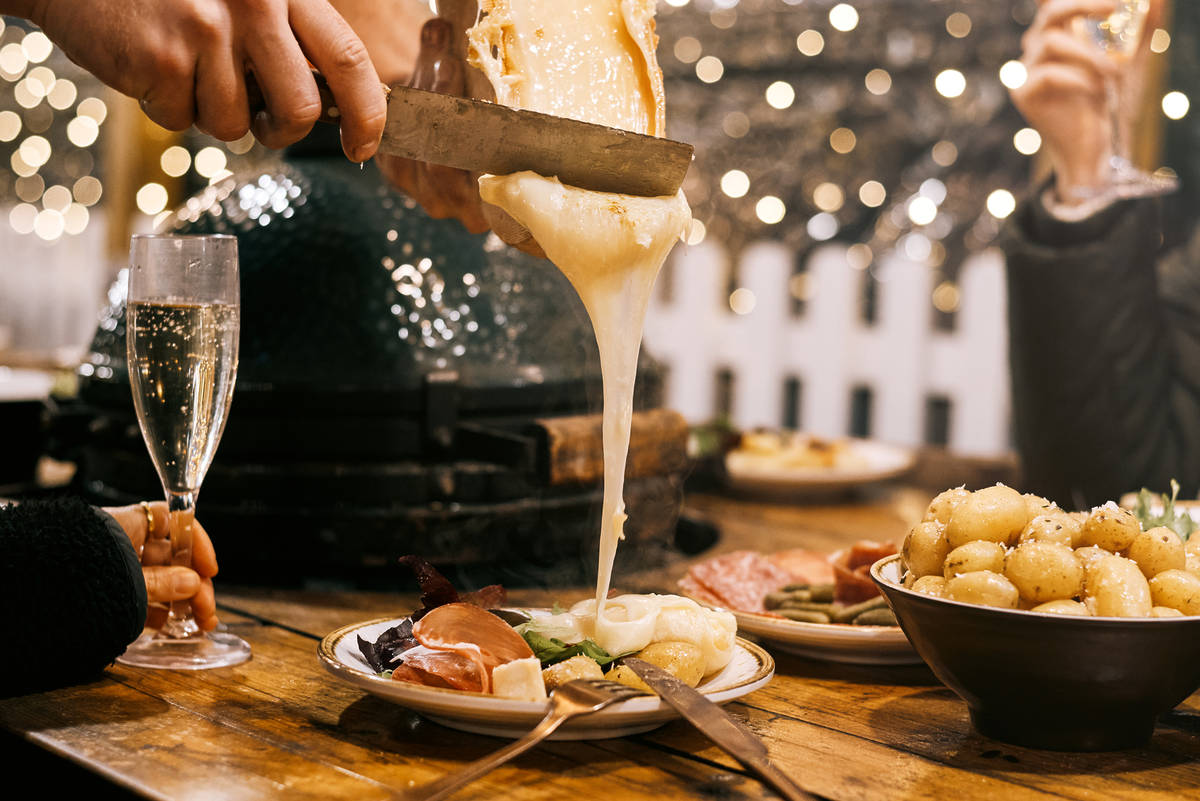 someone scraping oozy cheese off a wheel of raclette onto a waiting plate of food