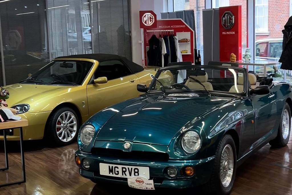 Un MG clásico verde azulado en primer plano con un MG deportivo dorado al fondo en un concesionario de coches.