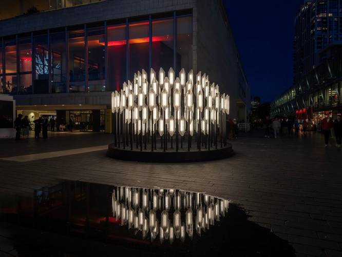 A striking outdoor light installation of a ring of glowing, upright crystalline shapes reflected in a puddle on a dark city square.