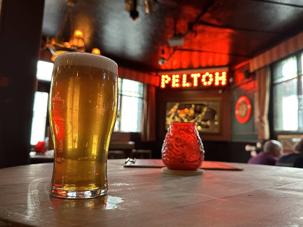 a pint of beer placed on a table in a pub, with a neon sign in the background reading 'PELTON;