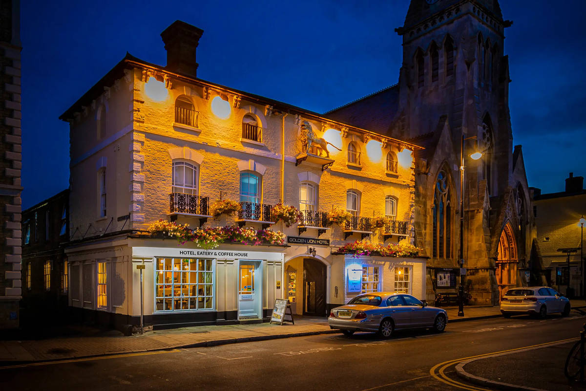 the exterior of the golden lion pub hotel at night, lit up softly with golden lights, next to a looming church building