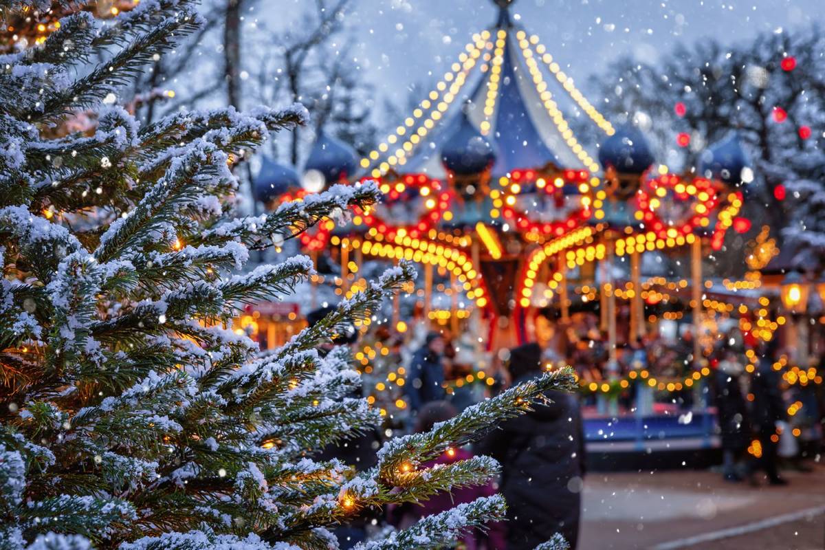 a snowy christmas tree in the foreground, with a merry-go-round lit up in the backgrund