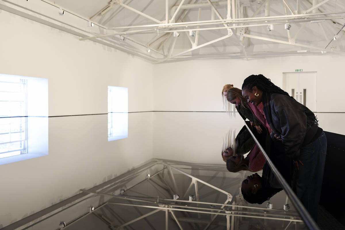people standing on a walkway in a room filled with oil, peering at their reflections in the still surface of the oil