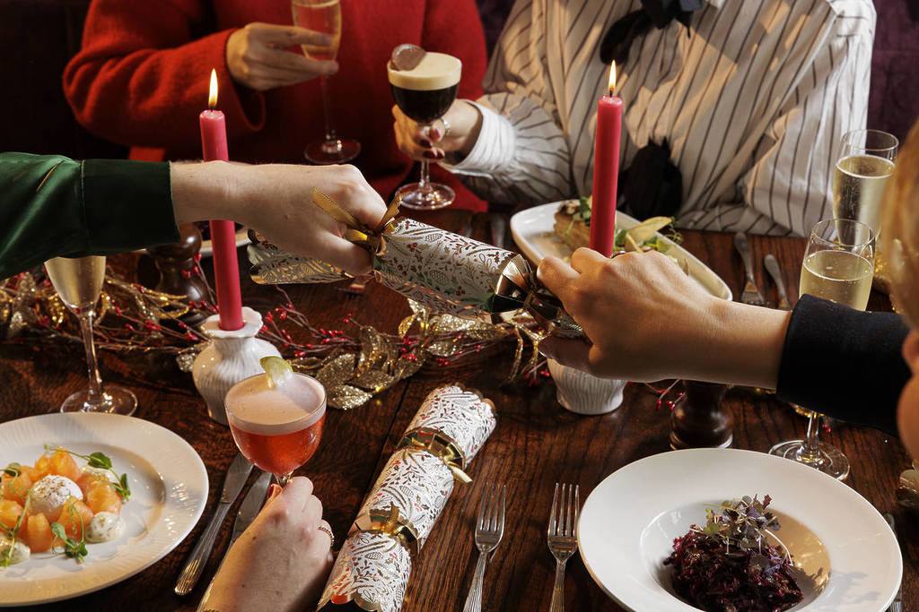 two hands pulling on a christmas cracker over a spread of christmas food, with two people in the background holding drinks