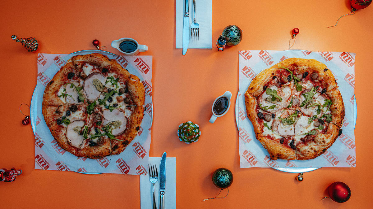 two pizzas on a table topped with turkey, chipolata sausage pieces, cranberry, and rocket, with a side of thick gravy dipping sauce