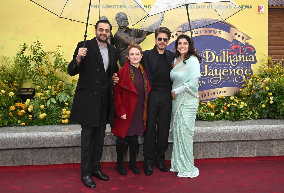 Akshaye Widhani, Ros Morgan, SRK & Kajol stood under umbrellas, in front of the newly unveiled bronze sculpture of bollywood actors from ddlj