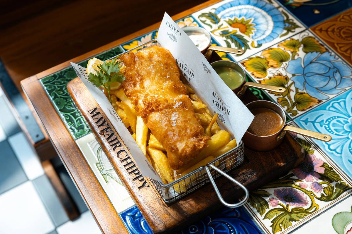 a whopping portion of fish and chips served in a fry basket atop a chopping board emblazoned 'mayfair chippy', alongside small pots of mushy peas and sauce