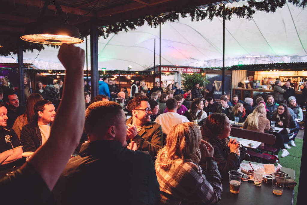 people sat at tables in a courtyard watching sports on a big screen, just out of shot