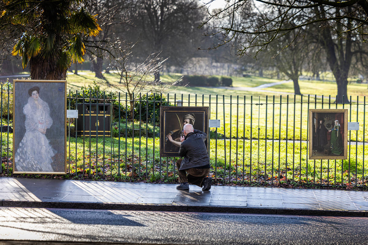someone walking past three famous paintings suspended on a fence by a park