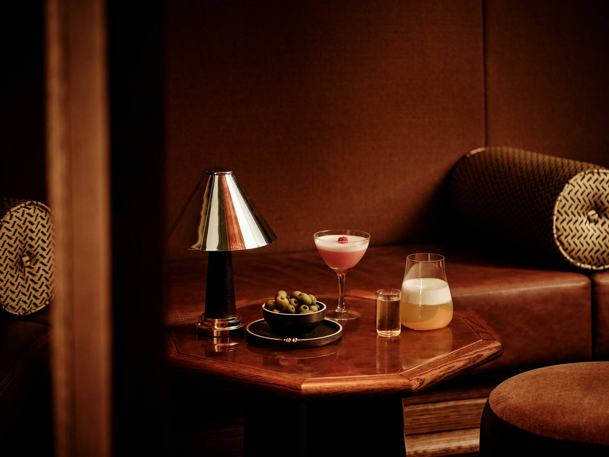 a low table in a softly-lit bar with a lamp, a bowl of olives, a red-hued cocktail in a martini glass, and a small glass of beer