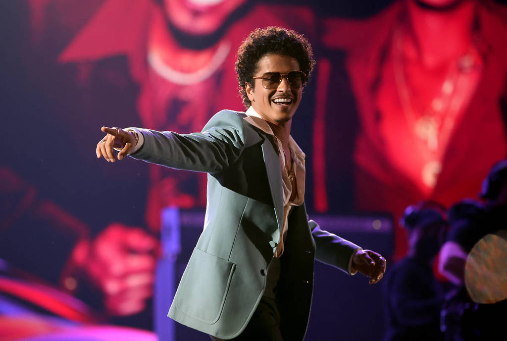 Bruno Mars con gafas de sol mirando y señalando hacia atrás.