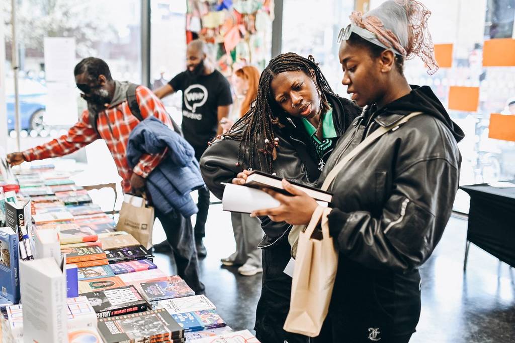 Two people examine a book with another reaching for a book within a selection of titles
