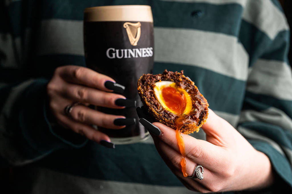 someone with long black painted nails in a striped rugby shirt holding a pint of guinness and a cut open 'guinness scotch egg' with a runny yolk