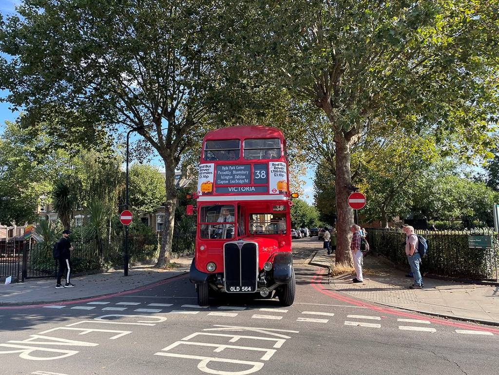 Ein alter Londoner Bus fährt aus einer Kreuzung heraus, neben ihm stehen Leute auf dem Bürgersteig.