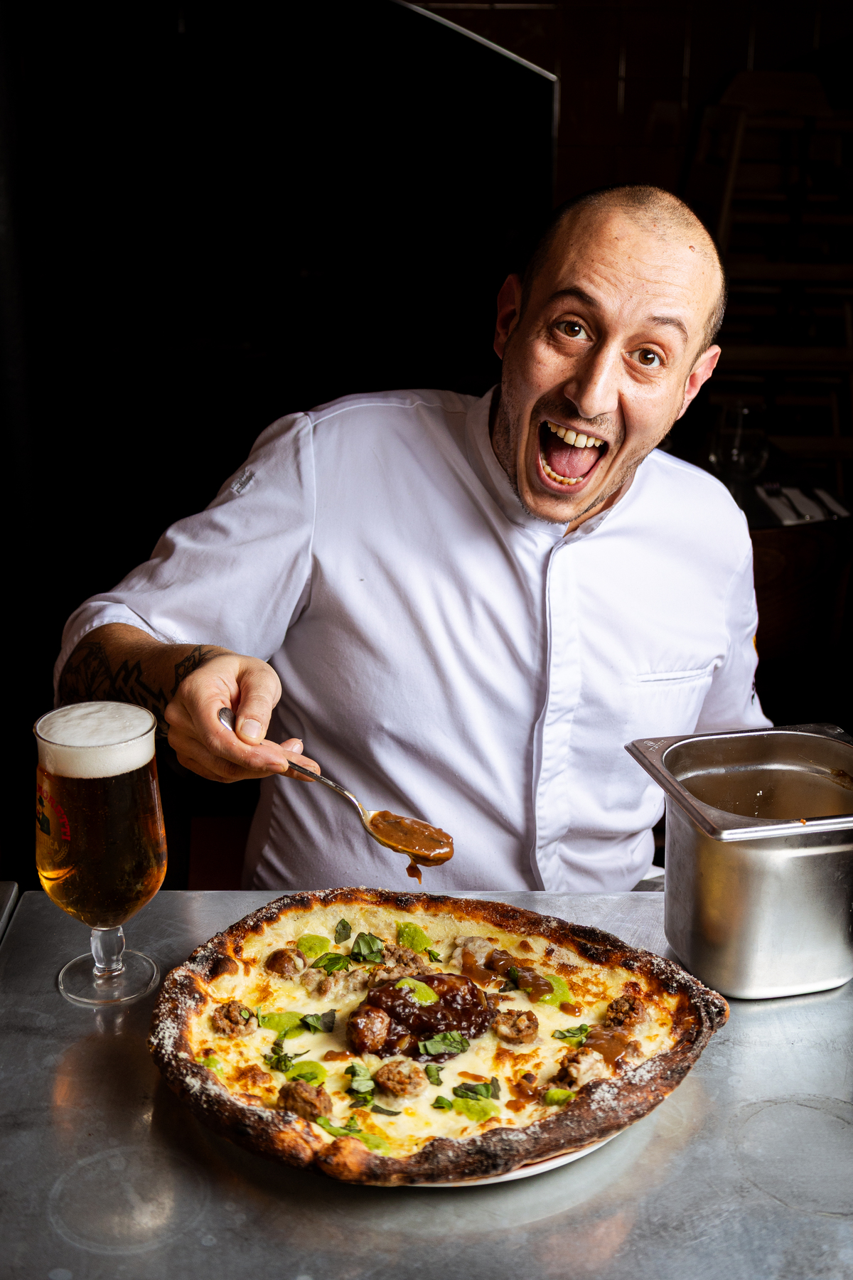 pizza chef Andrea Asciuti mugging form the camera as he dollops gravy onto a sausage and mash pizza, with a beer to the side